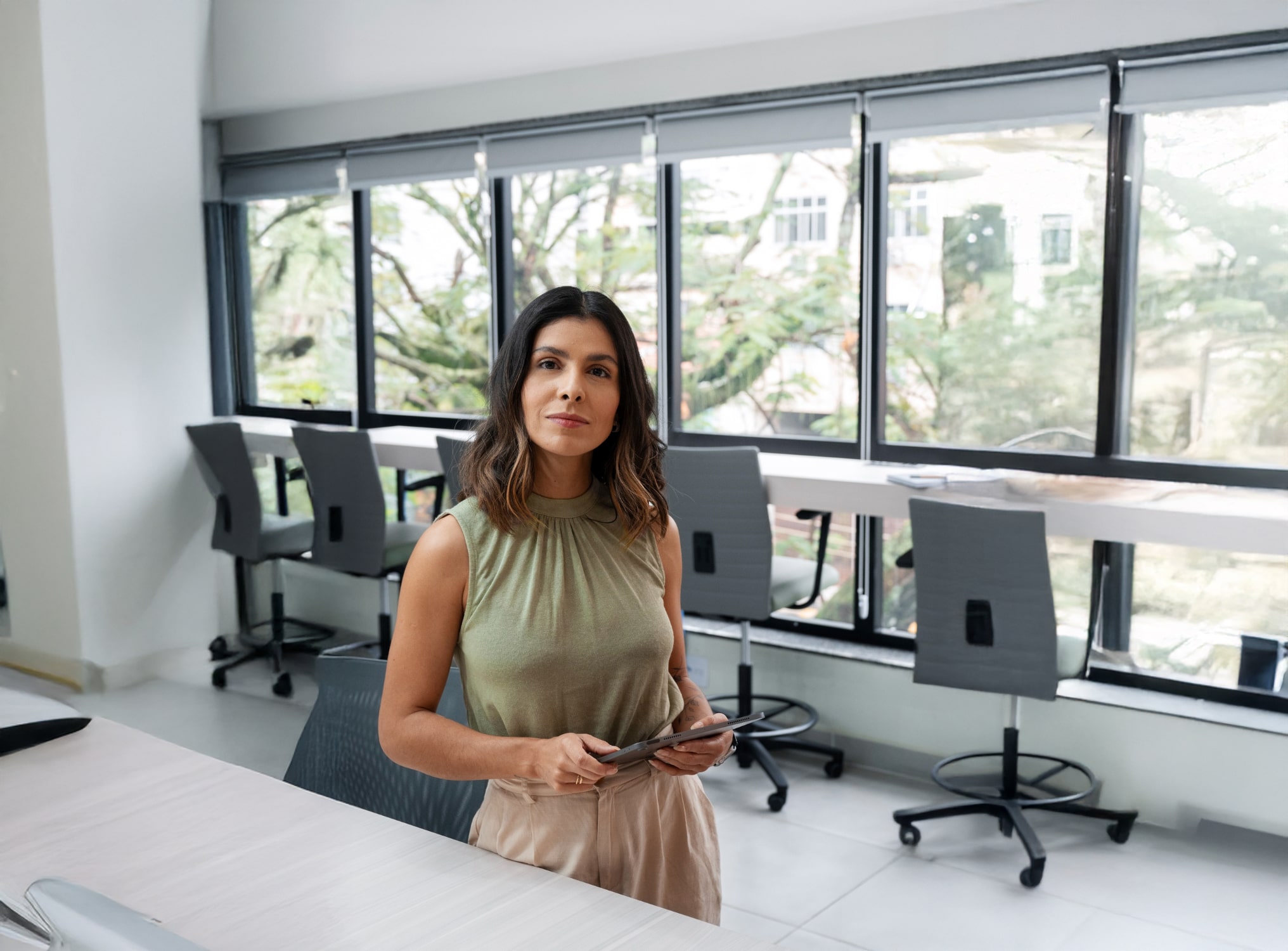 A woman with shoulder-length dark hair stands in a modern office, holding a tablet. She wears a sleeveless green top and beige pants. Large windows and empty chairs are visible in the background.