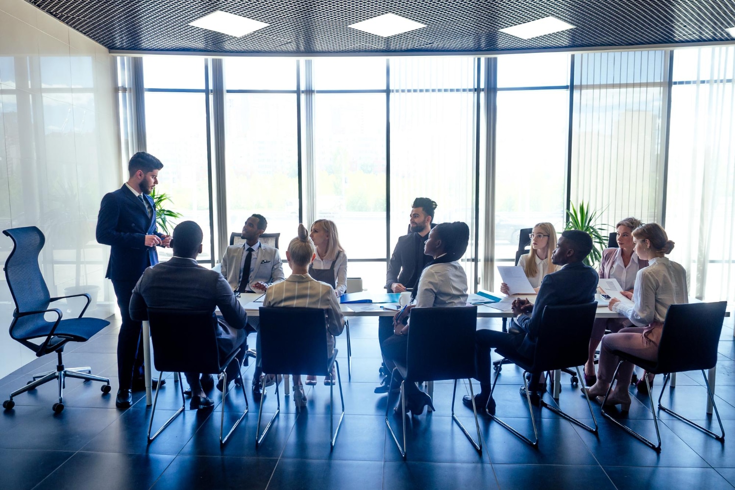 A group of business professionals sit around a conference table in a modern office, listening to a man standing and speaking. Large windows provide natural light, and there are plants in the background.