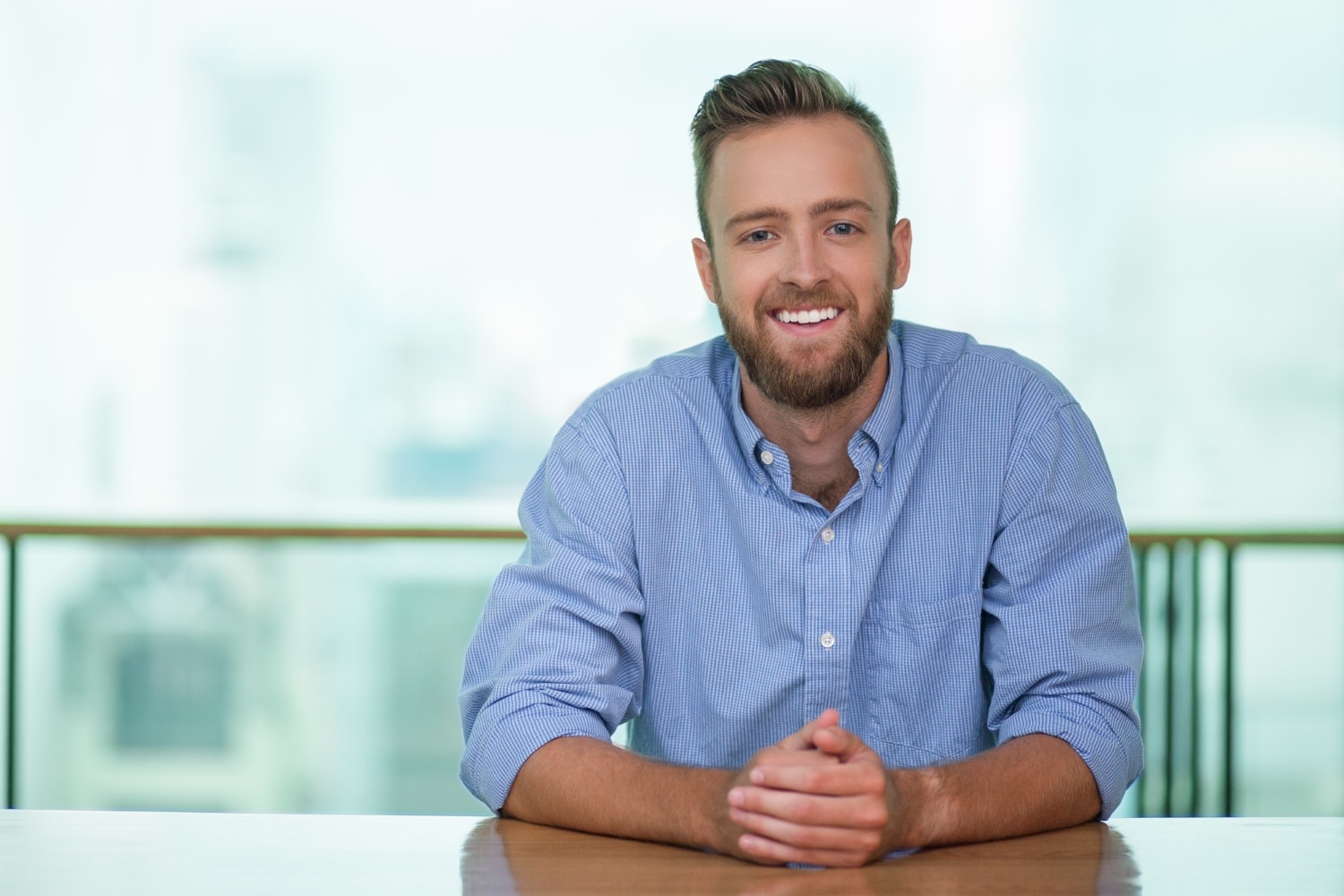 A man with a beard and short hair, wearing a light blue button-up shirt, sits at a table with his hands clasped and smiles at the camera. The background is blurred and bright.
