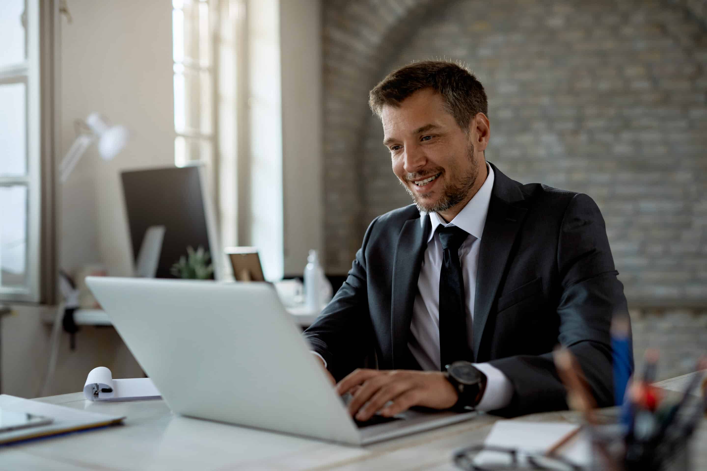 A man in a suit sits at a desk, smiling while working on a laptop in a bright, modern office with exposed brick walls and sunlight streaming through large windows.