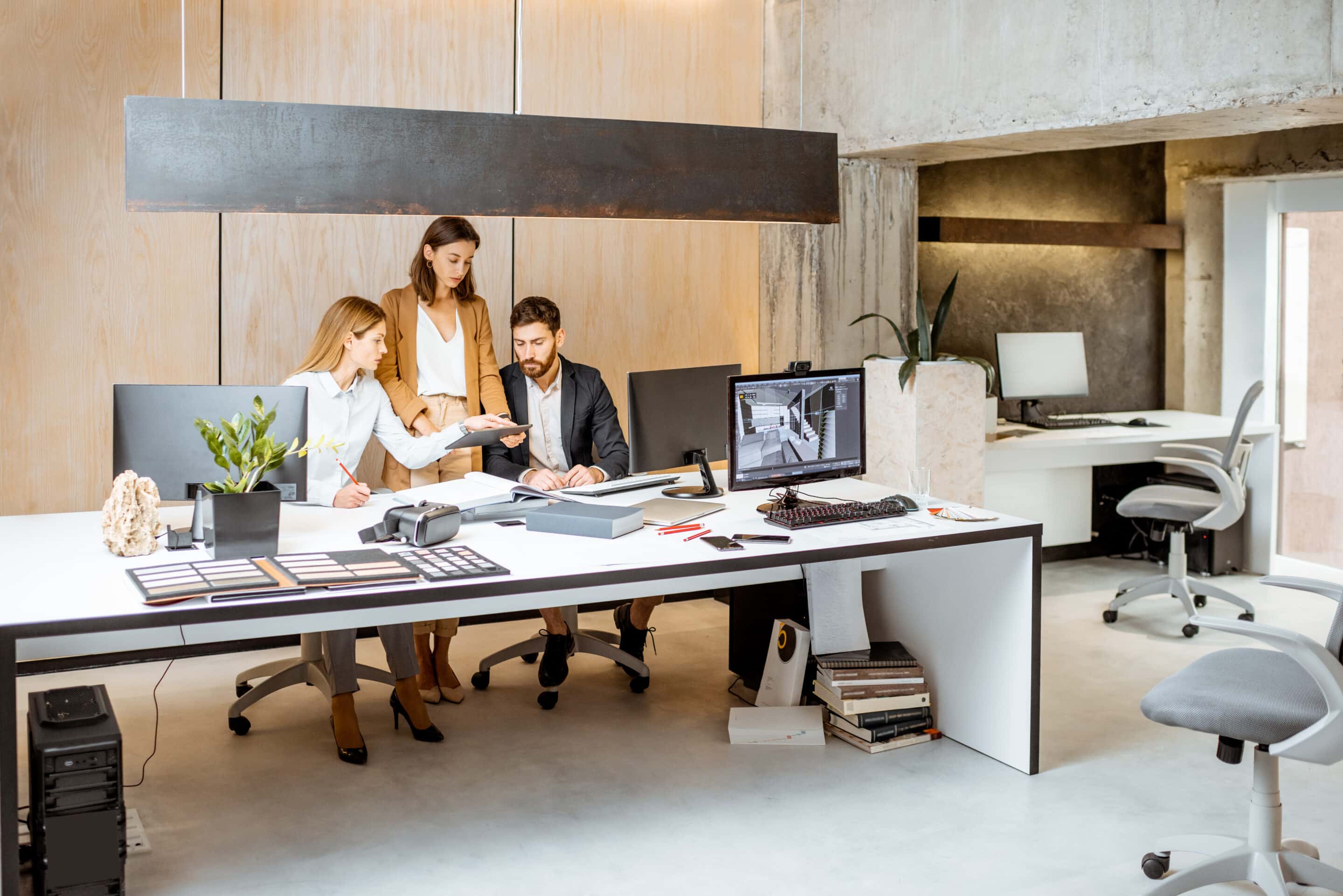 Two people in business attire shaking hands over a desk with a laptop, a small model house, a clipboard with documents, and a set of keys, symbolizing a real estate or business agreement.