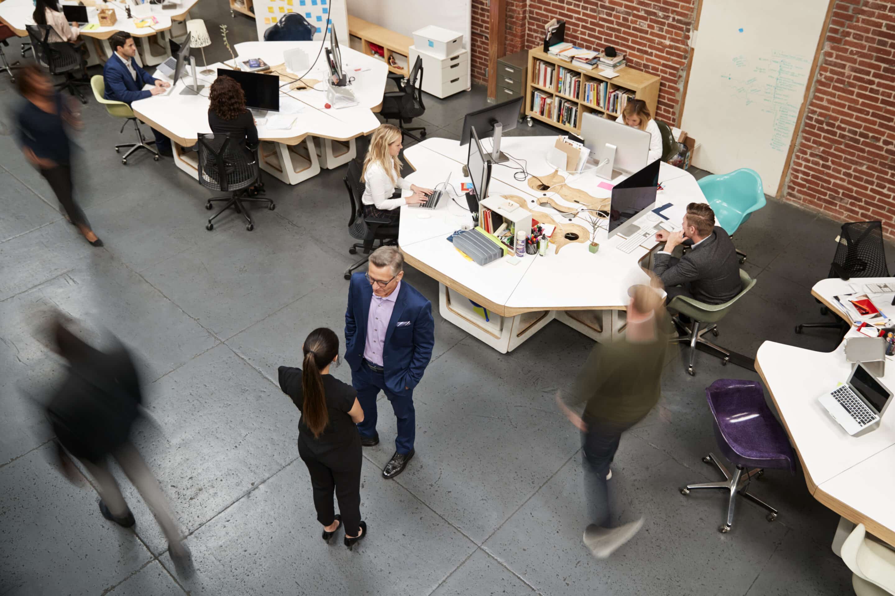 An overhead view of a modern office with people working at desks, some talking, and a few walking. Computers, papers, and office supplies are on desks, and there are exposed brick walls and shelves in the background.