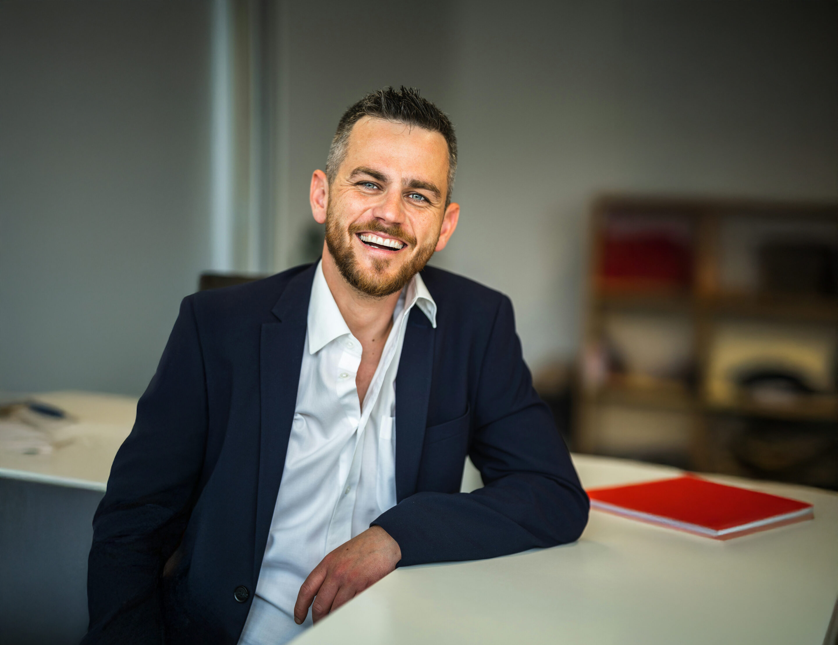 A man with short brown hair and a beard, wearing a dark suit and white shirt, sits at a desk smiling. There is a closed red notebook on the white desk, and shelves are blurred in the background.